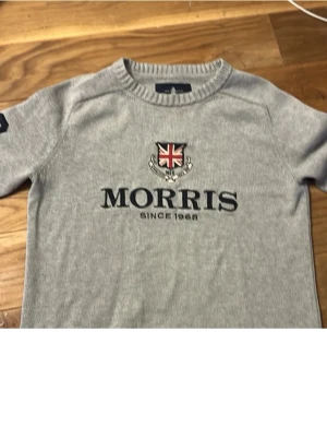 Grå stickad tröja från Morris - Snygg grå stickad tröja från Morris med broderad logga och brittisk flagga på bröstet. Rund hals och ribbade muddar ger en klassisk look. Perfekt för dig som gillar stilrena och tidlösa plagg. Passar bra på mig som är 175. 