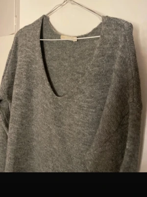 Grå stickad tröja med v-ringning 157 - Mysig grå stickad tröja från 157 i storlek S. Tröjan har en djup v-ringad hals och långa ärmar med ribbade muddar. Perfekt för kyliga dagar och enkel att matcha med jeans eller kjol. Materialet är mjukt och ger en avslappnad vibe.