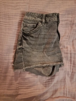 Grå jeansshorts från Lindex - Säljer ett par grå jeansshorts från Lindex med hög midja och råa, fransiga kanter. Shortsen har klassiska fem fickor, knapp och dragkedja framtill samt snygga detaljer på bakfickorna. Perfekta för varma dagar och chill stil.