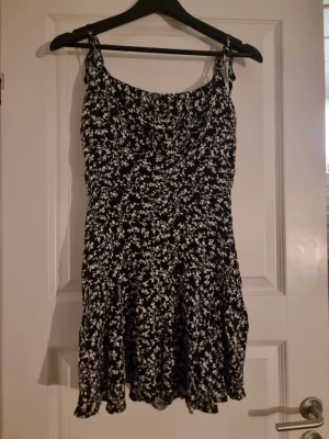 Svart blommig kortklänning med knyt - Supersöt svart klänning med vita blommor och knytbara axelband. Klänningen har en flowy kjol med volangkant och är ärmlös. Perfekt för varma dagar och har en lätt, luftig känsla. Materialet känns som tunn polyester eller bomull.