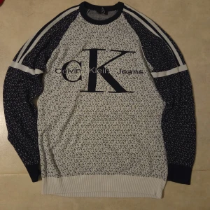 Mönstrad tröja från Calvin Klein Jeans - Säljer en snygg långärmad tröja från Calvin Klein Jeans med stort CK-tryck på bröstet. Tröjan är vit och mörkblå med ett småmönstrat print och vita ränder på ärmarna. Ribbad mudd vid hals, ärmslut och nederkant. Perfekt för dig som gillar streetwear.