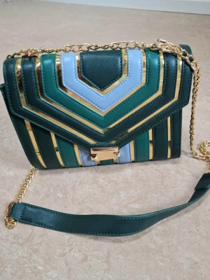 Blå grön axelväska med gulkedja - Very pretty bag with golden details and chain ✨️ no defects, except for none-noticable scratches on the clip (see picture - only if you look really close you can see it in real life). 1 compartment with zipper and 2 without. Bought second hand for 45€, but only used it once 🤗 Measurements: 24 cm broad, 19 cm high 👜