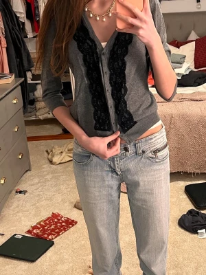 Grå kofta med svart spetsdetalj - Snygg grå kofta från H&M med svarta spetsdetaljer längs knappraden framtill. Koftan har trekvartsärm och är tillverkad i mjuk bomullsmix. Perfekt att styla med jeans eller kjol för en cool och feminin look.