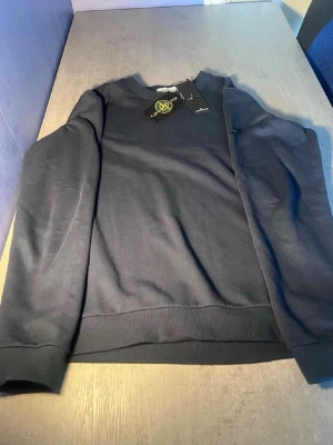 Mörkblå sweatshirt från Stone Island - Säljer en mörkblå sweatshirt från Stone Island med klassisk rund hals och ribbade muddar. Tröjan har den ikoniska Stone Island-loggan på ärmen och är tillverkad i mjuk bomull. Perfekt för dig som vill ha en stilren och enkel look.