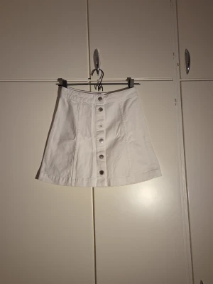 Vit kortkjol från H&M Divided - Säljer en vit kortkjol från H&M Divided i storlek 36. Kjolen är tillverkad i 100% bomull och har en A-linjeformad siluett med snygga silverfärgade knappar framtill. Perfekt för dig som gillar enkel och stilren design.