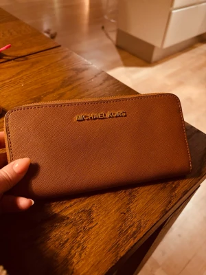 Brun plånbok från Michael Kors - Säljer en snygg brun plånbok/väska från Michael Kors. Plånboken har dragkedja runt om och flera fack för kort och sedlar. Insidan har diskret MK-mönster och är praktisk för vardagsgrejer.