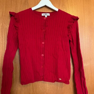 Röd kofta med volang från Hampton Republic - Snygg röd bomullskofta från Hampton Republic med kabelstickat mönster och volangdetaljer vid axlarna. Koftan är mycket sparsamt använd och saknar fläckar eller tecken på slitage.