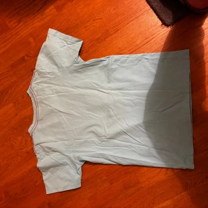 Ljusblå t-shirt med surftryck från Molo - Säljer en ljusblå t-shirt från Molo i storlek 152. T-shirten har ett stort färgglatt tryck med surfare och palmer på bröstet, samt en kontrastfärgad röd insida vid halsen. Materialet är mjuk bomull och modellen är klassiskt kortärmad.