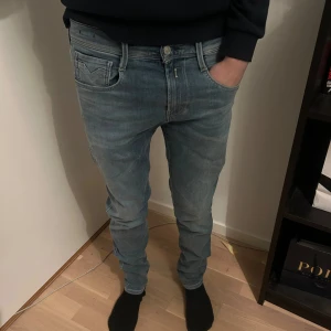 Replay Anbass ljusblå jeans slim fit - Säljer ett par Replay Anbass jeans i ljusblå tvätt med klassisk femficksdesign och snygga detaljer på bakfickorna. Jeansen har slim fit-passning och är tillverkade i mjukt stretchigt denim för skön komfort. Perfekt för dig som gillar stilrena och moderna jeans.