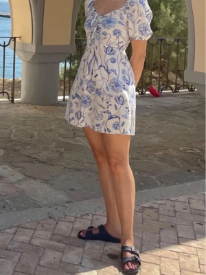 Vit och blå blommig kortklänning H&M - Supersöt kort klänning från H&M med puffärmar. Klänningen är vit med blått blommigt mönster och har snörning framtill. 