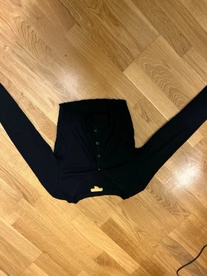 Svart ribbad kofta från Gina Tricot - Svart ribbad kofta från Gina Tricot med lång ärm och knappar framtill. Modellen är slim och har en klassisk rund halsringning. Perfekt att styla med jeans eller kjol för en enkel och snygg look.