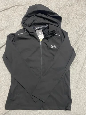 Svart hoodie från Under Armour M - Svart hoodie från Under Armour med dragkedja, huva och vita detaljer. Tillverkad i lätt syntetmaterial med logga på bröstet och Under Armour-text på ryggen. Passform är regular och materialet är andningsbart, perfekt för träning. Helt ny med lappar. Det var en julklapp till sonen  men för liten tyvärr.