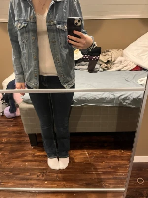 Klassisk blå jeansjacka med knappar - Säljer en klassisk blå jeansjacka med snygga metallknappar framtill och två bröstfickor med lock. Jackan har en rak passform och långa ärmar, perfekt att slänga på över en t-shirt. Materialet är robust denim och färgen är ljusblå.