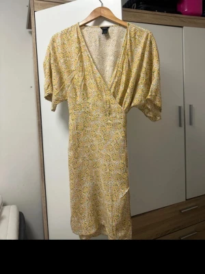 Blommig omlottklänning från Lindex - Säljer en söt kort omlottklänning från Lindex med blommigt mönster i gult, vitt och grönt. Klänningen har korta ärmar, v-ringad hals och knyt i midjan. Perfekt för varma dagar och har en luftig, skön känsla tack vare det tunna bomullsmaterialet.