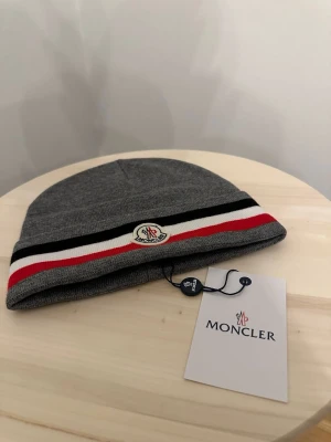 Grå Moncler mössa med ränder - Säljer en helt ny mössa oanvänd, Storlek One size Fraktar snabbt inom 24h efter köp.Ställ gärna din fråga.
