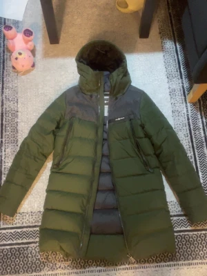 Grön lång pufferjacka med huva - Säljer en lång, mörkgrön pufferjacka från Sail racing med grå axelparti och huva. Jackan har två stora snedställda dragkedjefickor framtill och en markerad quiltad design. Perfekt för kalla dagar och har en clean streetstil.