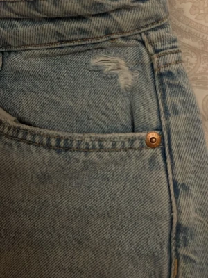 Ljusblå jeansshorts🩵 - Säljer ett par högmidjade ljusblå jeansshorts med fransiga kanter och slitna detaljer vid fickorna. Klassisk femficksmodell med knapp och dragkedja framtill. Perfekta för varma dagar och har en loose passform.