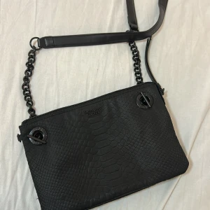 Svart axelväska från Victoria's Secret - Snygg svart axelväska från Victoria's Secret med reptilpräglat mönster på utsidan och kedjedetaljer på axelremmen. Insidan är fodrad med leopardmönster och har flera fack för bra organisering. Perfekt för dig som vill ha en cool och trendig väska.
