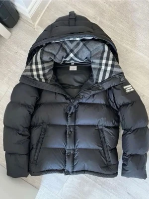 Svart pufferjacka från Burberry - Säljer en svart pufferjacka från Burberry med avtagbara ärmar så den kan användas som väst. Jackan har huva, dragkedja och knappar, samt klassiskt rutigt foder i huvan. Två fickor med dragkedja och Burberry-logga på ena ärmen. Perfekt för kalla dagar.