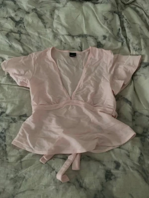 Ljusrosa omlottopp med knyt - Säljer en ljusrosa omlottopp från Gina Tricot med korta ärmar och v-ringad hals. Toppen har knytband i midjan och är tillverkad i mjuk bomull. Perfekt för dig som gillar feminina och stilrena plagg.