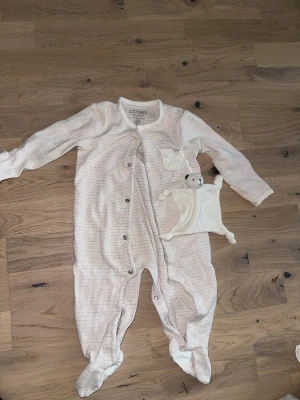 Randig babybody från Losan Losan Pyjamas med Snuttefilt nalle - Säljer en söt babybody från Losan i ljusbeige med tunna rosa och grå ränder. Västen har långa ärmar, tryckknappar framtill och en liten ficka med ett mjukt nalle-gosedjur. Perfekt för mysiga dagar hemma.Söt pyjamas med tillhörande snuttefilt. Pyjamasen har en ljus färg med tunna ränder och knäpps med tryckknappar framtill. Den har även en liten ficka på bröstet.