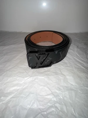 svart skinnbälte - Säljer ett bälte i svart skinn med klassisk logga på spännet. Bältet har diskret mönster och snygga detaljer i svart metall. Insidan är ljusbrun med präglad märkning och kontrastsömmar. Perfekt accessoar för att lyfta din outfit.