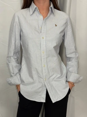 Ralph Lauren skjorta - Säljer en klassisk vit och ljusblå randig skjorta från Ralph Lauren med broderad logga på bröstet. Skjortan har lång ärm, knappar framtill och en snygg krage. Perfekt för dig som gillar stilrena och tidlösa plagg. Det är en dam skjorta och är i storlek XS.