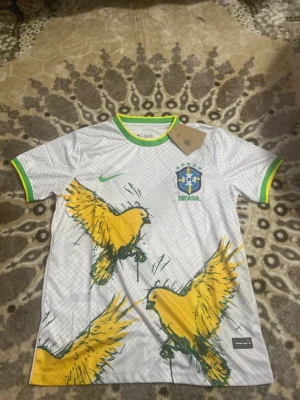 Vit Brasil Nike fotbollströja - Säljer en vit Brasilien fotbollströja från Nike med gröna och gula detaljer på krage och ärmslut. Framsidan har grafiskt mönster och stora gula fåglar, samt Brasilien-emblem och Nike-logga. Materialet är lätt polyester som andas.