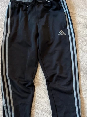 Adidas strl 140 - Bilden visar ett par svarta Adidas träningsbyxor med grå ränder längs sidorna och Adidas-logga på låret. Byxorna har snörning i midjan och är tillverkade i ett syntetmaterial.