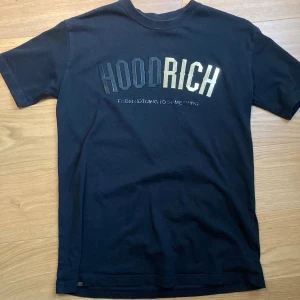 Svart HOODRICH t-shirt XS - Svart t-shirt från HOODRICH i storlek XS med tryck i svart och guld på bröstet där det står 'HOODRICH' och 'From nothing to something'. Klassisk rund hals och korta ärmar. Mjuk bomullskvalitet som känns skön mot huden.