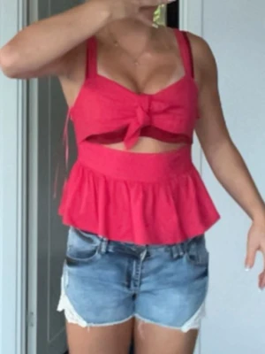 Röd volangtop med rosett och cutout - Säljer en röd topp med breda axelband, volang nedtill och snygg cutout under bysten. Toppen har en stor rosett framtill och är croppad i modellen. Perfekt för sommarens alla häng och har en feminin vibe med sin volangkant och knytning. SLUTSÅLD PÅ ZARA