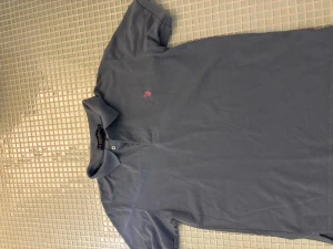 Blå piké Polo - Säljer en klassisk blå pikétröja från Ralph U.SN med orange broderad logga på bröstet. Stilig piké med en unik och ögonfångande färg så man sticker ut. Nice med linneshorts eller linnebyxor. Legat i garderob länge och därmed lite skrynklig men går att lösa enkelt bara att stryka en gång. Storlek S/M