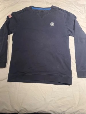 Mörkblå Sail Racing sweatshirt USA 77 - Mörkblå sweatshirt från Sail Racing med broderad amerikansk flagga på ärmen och tryck 'USA 77' på ryggen. Rund hals, långärmad och mjukt bomullsmaterial. Sail Racing Performance Technology-märke på bröstet. Perfekt för dig som gillar sportig och avslappnad stil.