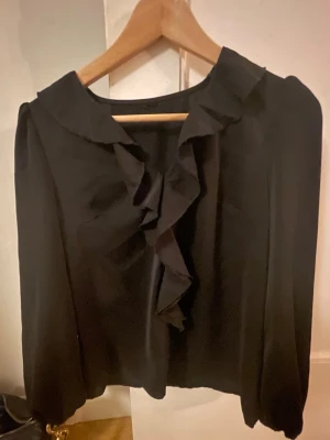 Svart blus  - Fin svart blus med volanger framtill. Tröjan är i storlek XS men fungerar också i S. Säljer eftersom den inte kommer till användning hos mig! Den är helt oanvänd. 