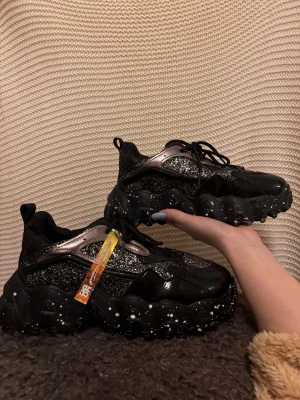 Svarta sneakers  - Säljer ett par svarta sneakers med glittriga metallic-detaljer, skorna är aldrig använda. Storlek 40 men passar som 39. Orginal pris: 660kr