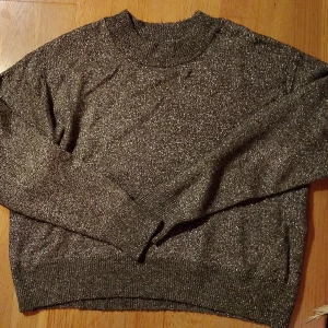 Glittrig mörkgrå stickad tröja - Snygg stickad tröja i mörkgrått med glittriga inslag. Tröjan har en rund hals och långa ärmar, perfekt för att ge din outfit lite extra shine. Ribbstickade muddar vid hals, ärmslut och nederkant. 