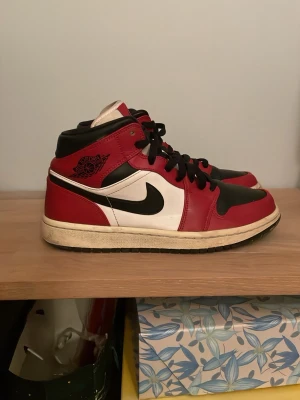 Nike Air Jordan 1 Mid röd/svart/vit - Säljer ett par Nike Air Jordan 1 Mid sneakers i klassisk röd, svart och vit färg. Skorna har snörning, rund tå och platt sula. Ikonisk Air Jordan-logga på sidan och svart swoosh. Ordenarie priss på nya 1800-3000kr 