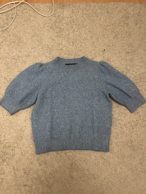 Blå stickad tröja Vero Moda S - Mysig blå stickad tröja från Vero Moda i storlek S. Tröjan har puffiga korta ärmar och ribbade muddar vid hals, ärmslut och nederkant. Perfekt för att ge din outfit en chill och trendig vibe. Färgen är melerad blå och materialet känns mjukt och skönt.