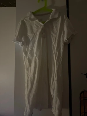 Vit kortklänning med krage från Nike - Säljer en vit kortklänning från Nike med klassisk pikékrage och tre knappar framtill. Klänningen har korta ärmar och är gjord i mjuk bomull, perfekt för en sportig och avslappnad look. Diskret Nike-logga broderad på bröstet.