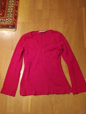 Rosa långärmad blus med broderi - Säljer en snygg rosa blus med lång ärm och v-ringad hals. Blusen har fina broderade blommor framtill som ger en extra detalj. Materialet känns mjukt och skönt, perfekt för dig som gillar färg och vill sticka ut lite.