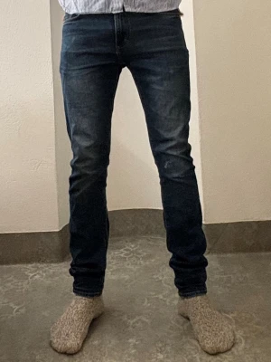 Mörkblå slim fit jeans från East West - Säljer ett par mörkblå slim fit jeans från East West. Jeansen har klassisk femficksdesign, raka ben och snygg tvätt med lätt slitning. Materialet är tåligt jeans-tyg i bomull. Perfekta för dig som gillar en smalare passform och stilren look. Modellen är 184 och väger 72 lite tighta i midjan. De har en snygg slitningadetalj som jag har gjort själv. 