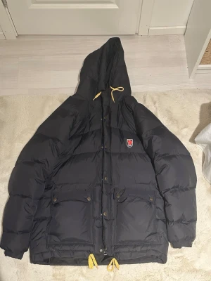 Svart dunjacka från Fjällräven - Svart dunjacka från Fjällräven med gul snörning i huvan och nederkant, stor huva och två stora fickor med knappar. Jackan har broderad logga på bröstet och grov dragkedja med metallringar. Perfekt för kalla dagar och riktigt snygg streetstil.