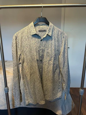 Blommig skjorta från Rocha John Rocha - Snygg långärmad skjorta från Rocha John Rocha med blommigt mönster i blått, vitt och små röda detaljer. Skjortan har klassisk krage, knappar framtill och är gjord i mjuk bomull. Perfekt för dig som gillar stilrena men ändå unika plagg.