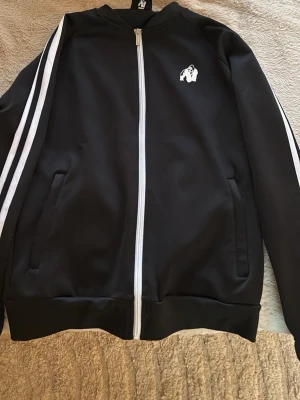 Svart zip tröja med vita ränder - Snygg svart ziptröja från Gorilla Wear med vita ränder längs ärmarna och vit dragkedja framtill. Jackan har ett stilrent gorilla-tryck på bröstet och två fickor på sidorna. Perfekt för dig som gillar sportig och clean stil.