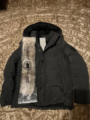 Canada Goose Wyndham Parka  - Säljer en mörkgrå Wyndham Parka från Canada Goose black label med huva och avtagbar pälsdetalj. Jackan har broderad logga på ärmen, dragkedja och knappar framtill. Perfekt för kalla vinterdagar och har en clean look.