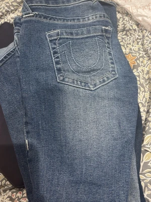 True Religion blå jeans, storlek 26 - Säljer ett par snygga blå jeans från True Religion i storlek 26. Klassisk femficksmodell med ikonisk U-söm på bakfickan och silverfärgade detaljer