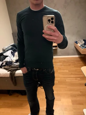 Mörkgrön stickad tröja från J.Lindeberg - Snygg mörkgrön stickad tröja från J.Lindeberg med rund hals och ribbade muddar. Perfekt passform som sitter normalt och långa ärmar. Stilren design utan mönster, passar dig som gillar enkelhet och kvalitet.