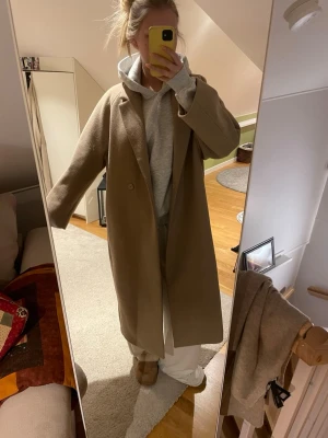 Beige lång kappa med klassisk krage - Säljer en stilren och lång beige kappa med klassisk krage och knäppning framtill. Kappan har en rak passform och är perfekt för dig som gillar en clean och tidlös look. Materialet känns mjukt och ger en lyxig känsla. Passar perfekt till lager-på-lager-stil under höst och vinter.