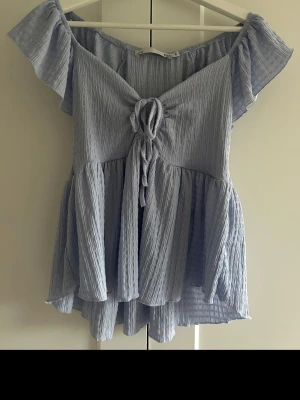 Topp - Supersöt ljusblå topp med knyt framtill. Blusen har offshoulder och korta volangärmar, samt ett flowigt fall nedtill.