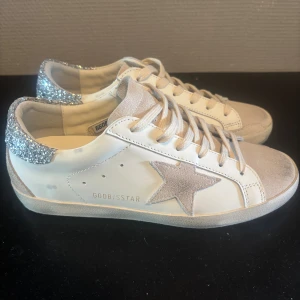 Golden Goose Superstar sneakers med glitter - Golden Goose Superstar sneakers i vitt skinn med beige mockadetaljer och en ikonisk stjärna på sidan. Baktill har de en silvrig glittrig häl som verkligen poppar. Klassisk snörning och platt sula. Kommer med originalpåse.